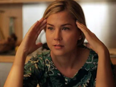 Woman with blond hair in a patterned green shirt looks contemplative, resting her hands on her temples.