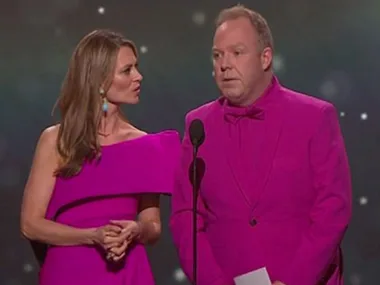 Two people in matching bright pink outfits stand on stage, one holding a microphone.