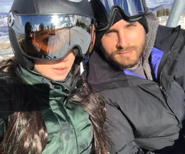 Two people in ski gear taking a selfie on a snowy mountain, one wearing reflective goggles.