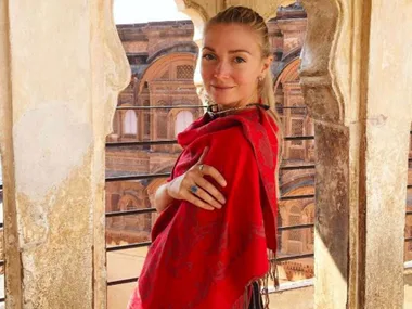 A woman in a red shawl stands in a historic stone structure with intricate architecture visible in the background.