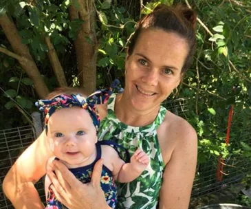 Mom smiling, holding baby in floral outfits, outdoors with greenery.
