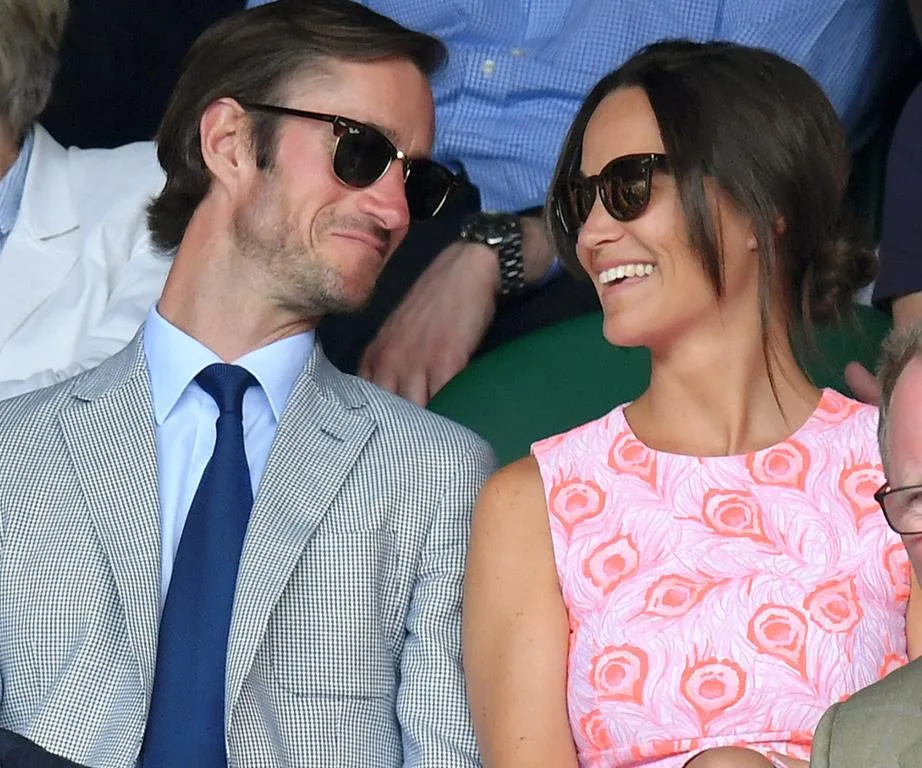 Couple wearing sunglasses and smiling at a sports event, seated among other spectators.