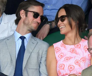 Couple wearing sunglasses and smiling at a sports event, seated among other spectators.