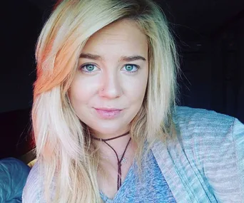 A woman with long blonde hair and a nose ring, wearing a necklace, looking at the camera in a softly lit room.