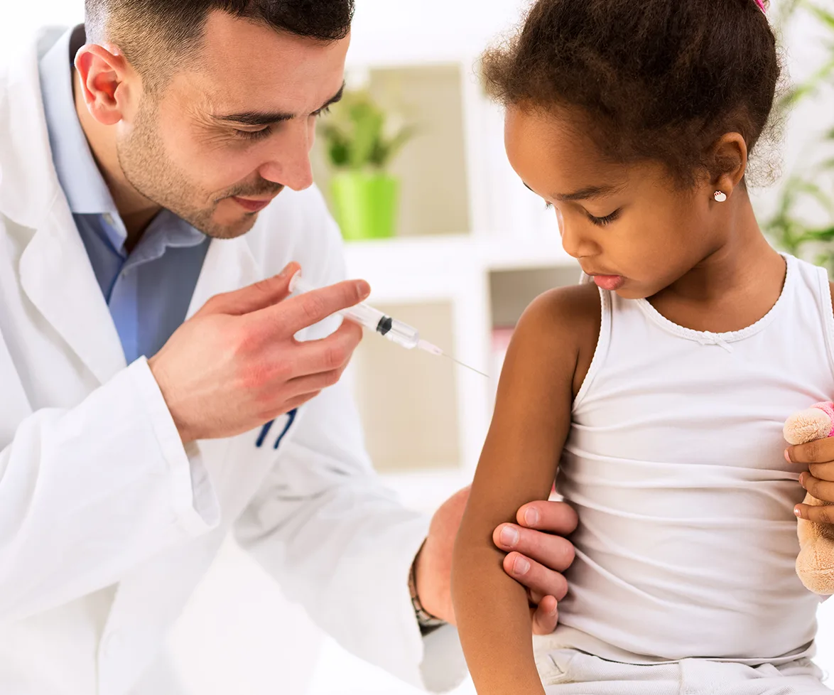 Doctor in white coat administers vaccine to young girl holding a teddy bear, in a bright room.
