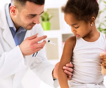 Doctor in white coat administers vaccine to young girl holding a teddy bear, in a bright room.