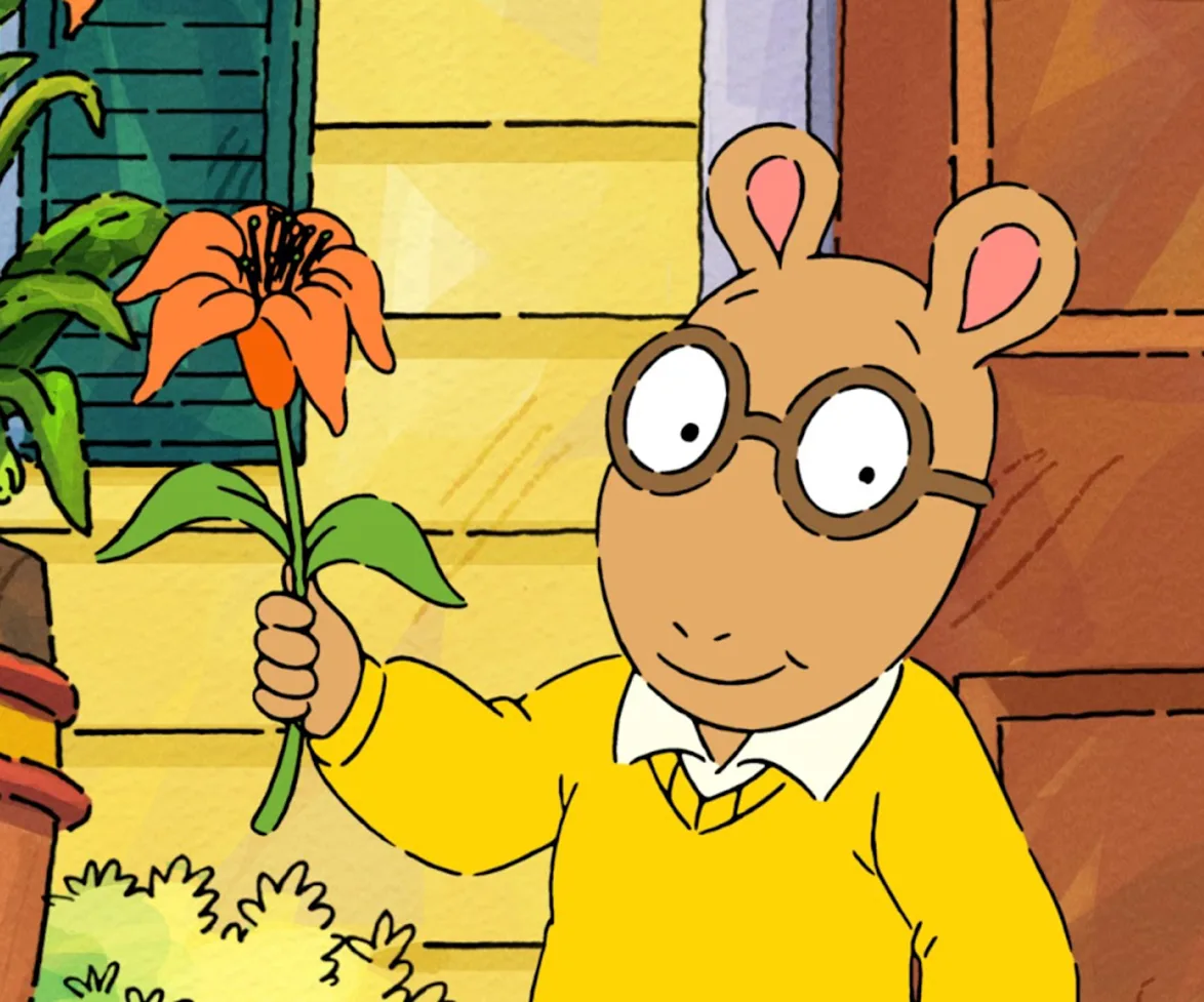 Arthur holding an orange flower in front of a house, wearing glasses and a yellow sweater.