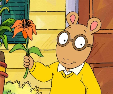 Arthur holding an orange flower in front of a house, wearing glasses and a yellow sweater.