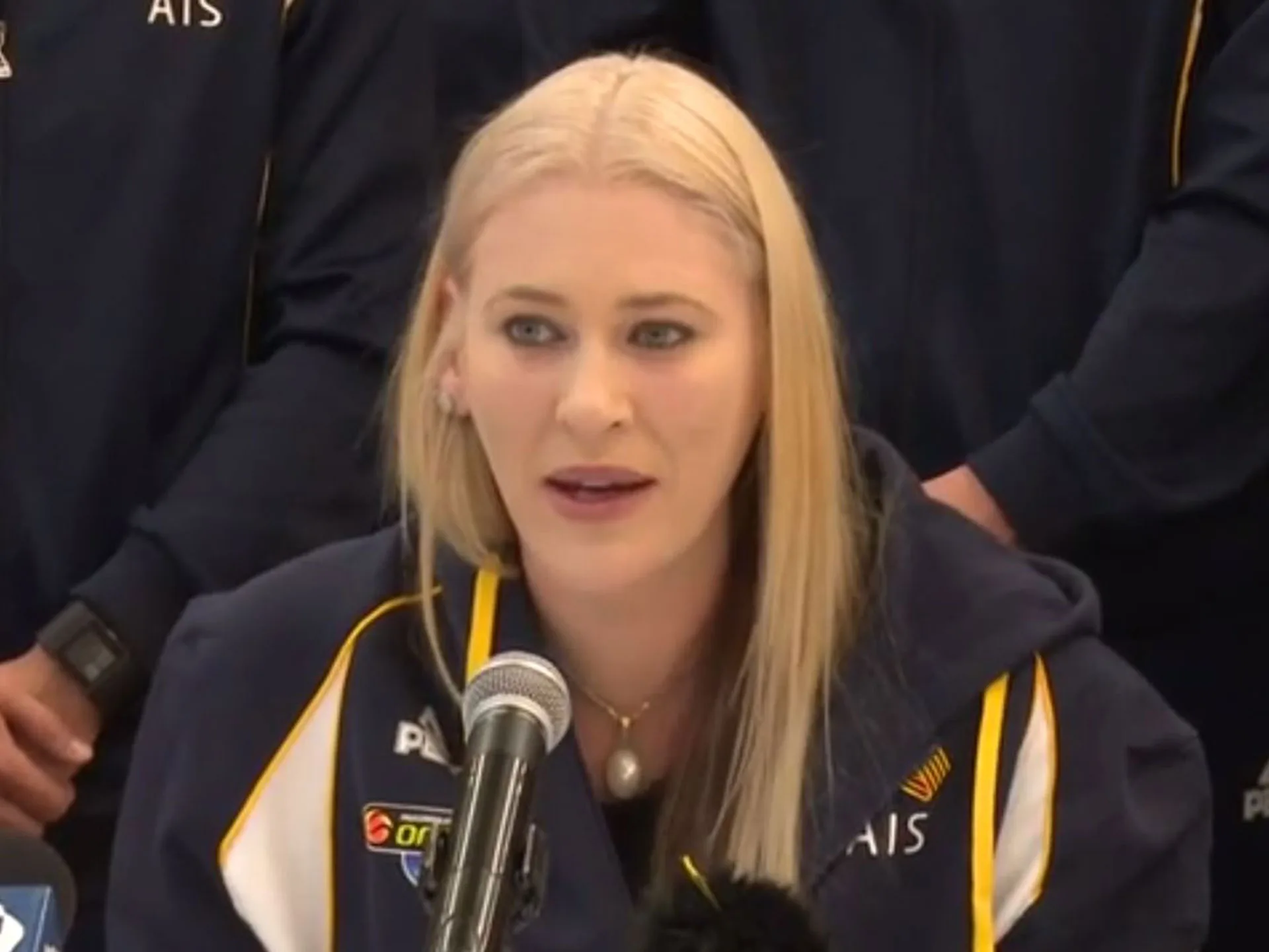 A person with blonde hair speaks at a microphone, wearing a sports jacket with "AIS" on it.