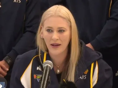 A person with blonde hair speaks at a microphone, wearing a sports jacket with "AIS" on it.