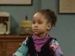 Young girl in colorful clothes shrugging with a confused expression indoors.