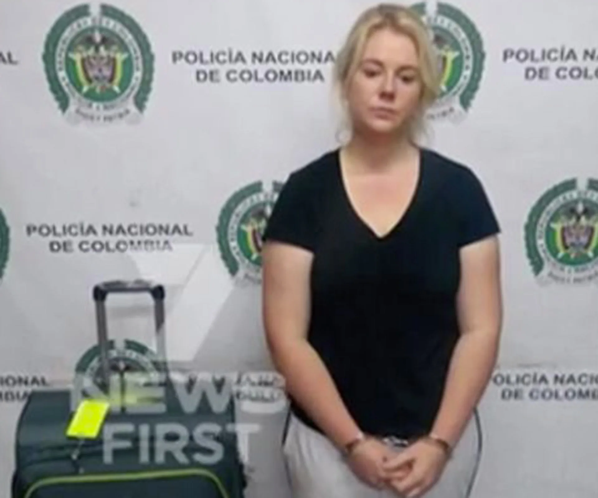 Woman standing with suitcase, against a wall with "Policia Nacional de Colombia" logos.