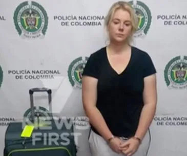 Woman standing with suitcase, against a wall with "Policia Nacional de Colombia" logos.