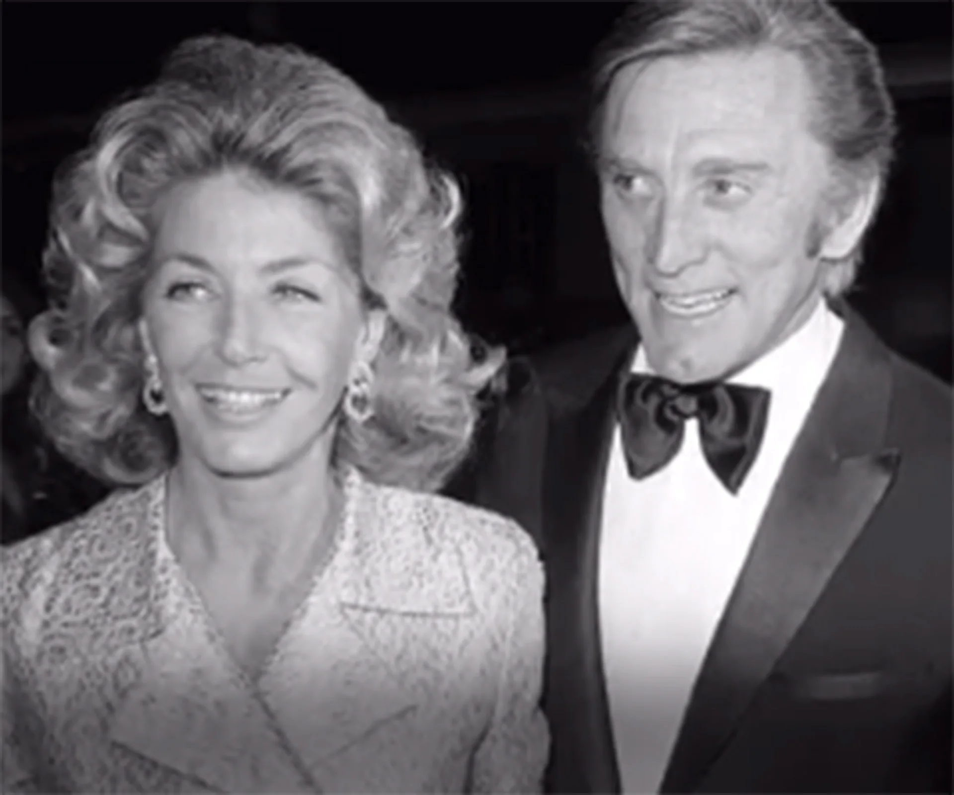 A well-dressed couple, the man in a tuxedo, the woman with styled hair and earrings, smiling together.