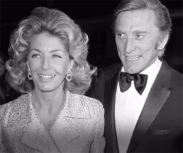 A well-dressed couple, the man in a tuxedo, the woman with styled hair and earrings, smiling together.