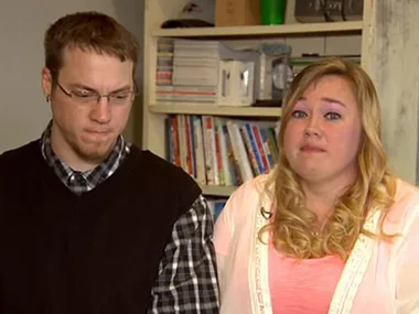 Man and woman sitting indoors, both appear serious, with a bookshelf in the background.
