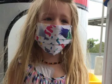 Young girl with long blonde hair wearing a colorful patterned mask, standing outside near a playground structure.