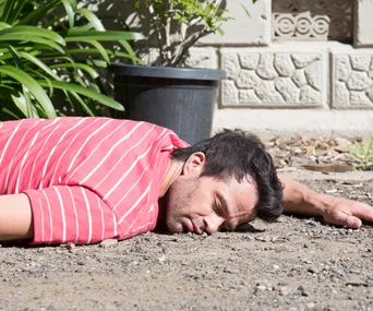 Man in red striped shirt lying face-down on gravel, next to potted plant, outdoors.