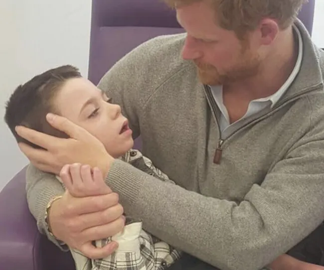Man in grey sweater holds a young boy tenderly in his lap, gazing at him with care in a seated setting.