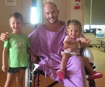 Man in wheelchair wearing hospital gown, smiling with two young children beside him and on his lap.