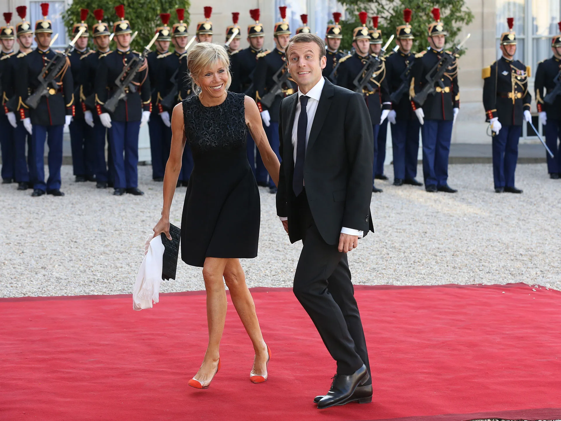 Couple walking on a red carpet, dressed formally, with a military guard of honor in the background.
