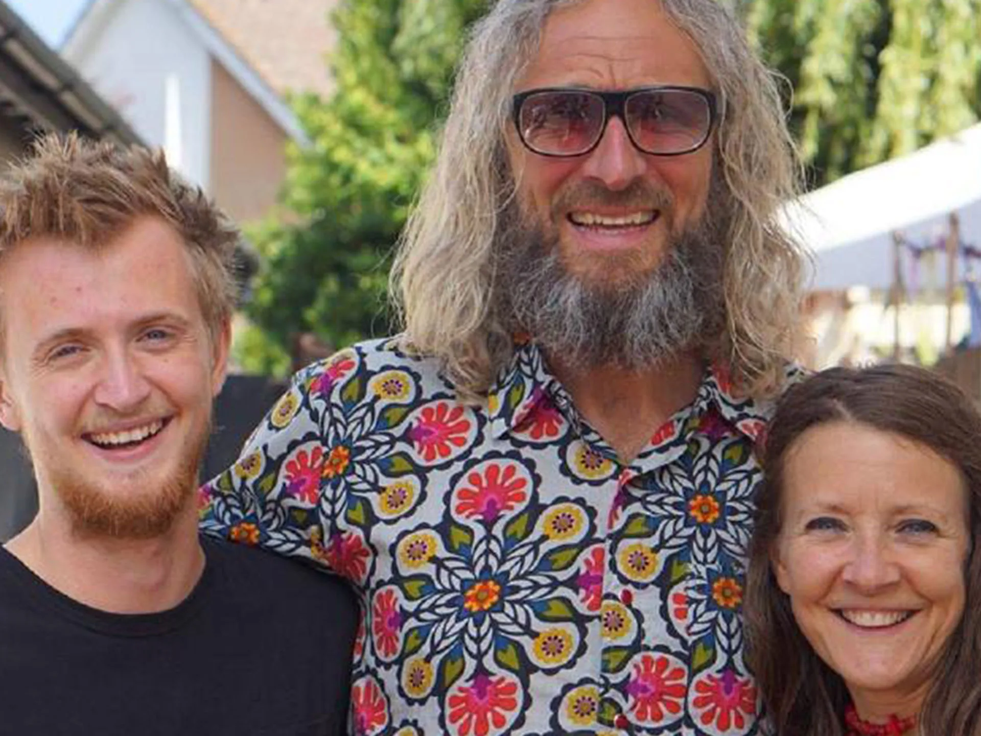 A man with long hair and glasses stands between a young man and a smiling woman outdoors.