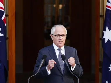 Person in a suit speaks at a podium with microphones, standing between two flags.