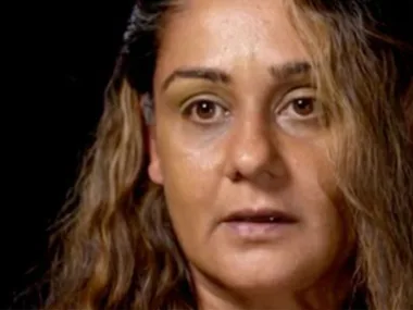 A woman with long brown hair and a serious expression against a dark background.