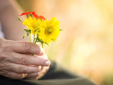 Hand holding a small bouquet of yellow and red flowers against a soft blurred background.
