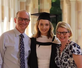 A happy graduate in a black cap and gown stands between two smiling adults, possibly parents, in a sunlit hall.