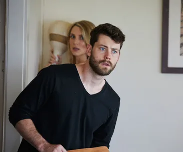 Man looking concerned in a room, woman in the background holding a hat.