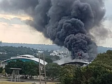 Thick black smoke rises from a fire at a building in Sydney Olympic Park, surrounded by trees and other structures.