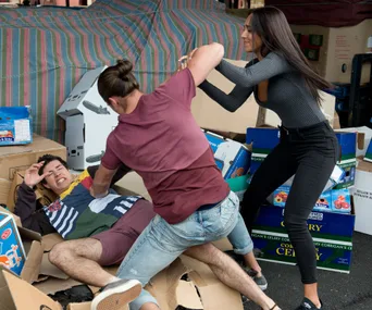 A man is on the ground among cardboard boxes, defending himself as two others appear to confront him aggressively.