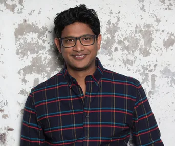 A person in glasses smiling, wearing a plaid shirt, stands in front of a textured wall.