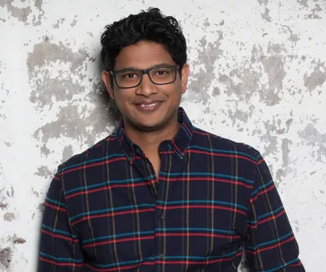 A person in glasses smiling, wearing a plaid shirt, stands in front of a textured wall.