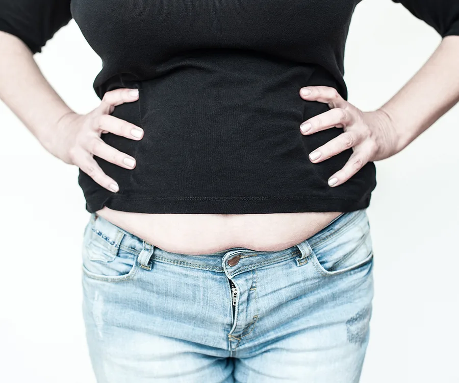 A woman in a black top and jeans showing her belly with her hands on her hips.