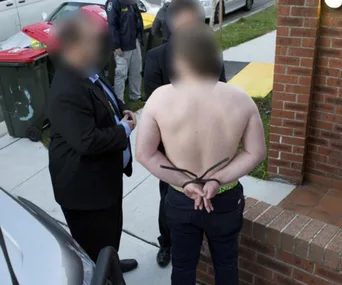 A shirtless man with hands tied behind, surrounded by blurred figures, possibly law enforcement, near a brick wall.