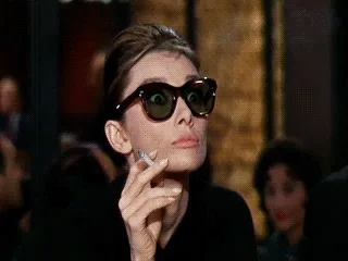 A woman with dark sunglasses holds a cigarette, looking to the side with a poised expression.