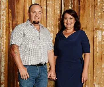 A couple holding hands, standing in front of a rustic wooden background, smiling at the camera.