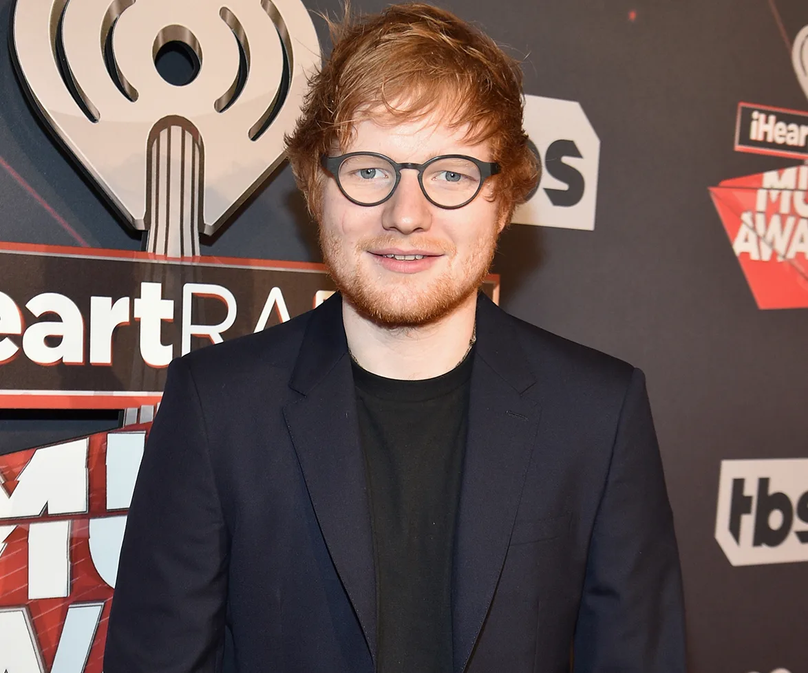 A man with glasses and red hair in a suit at a music awards event, standing in front of a logo backdrop.