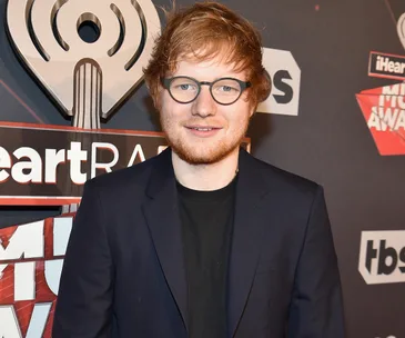 A man with glasses and red hair in a suit at a music awards event, standing in front of a logo backdrop.