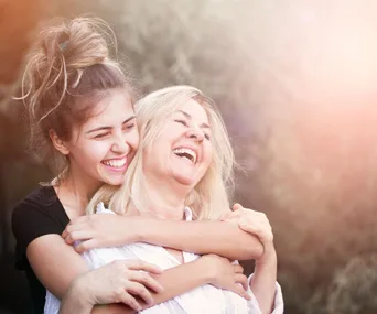 A young woman hugs an older woman from behind, both laughing joyfully in an outdoor setting.