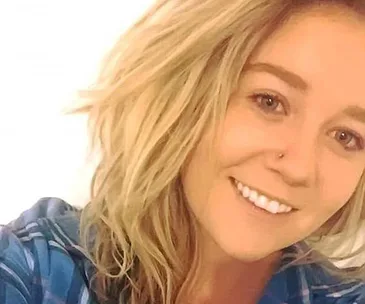 A woman with blonde hair and a nose piercing smiles in a selfie wearing a blue plaid shirt.