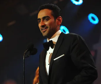 A person in a tuxedo speaks at a microphone on stage with bright lights in the background.