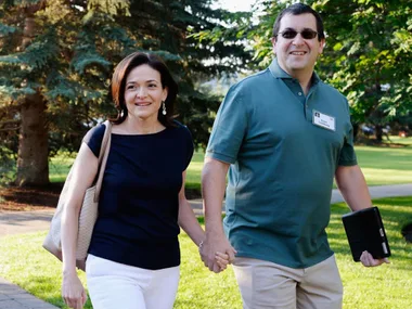 A smiling couple walks hand-in-hand outdoors, with trees and grass in the background.