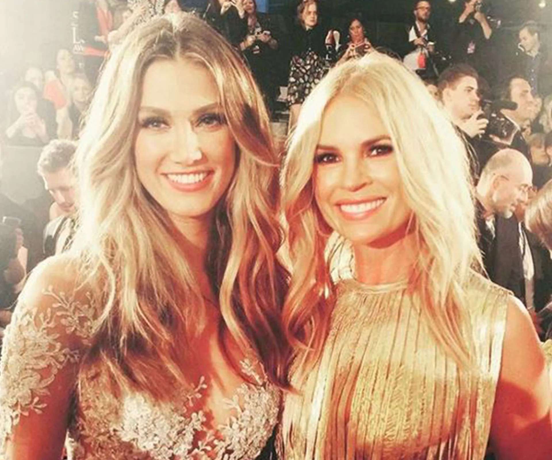 Two women smiling at an event, surrounded by an audience and photographers.