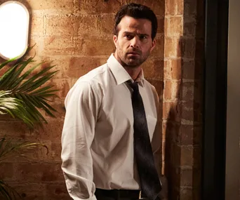 Man in a white shirt and tie stands thoughtfully against a brick wall with a plant nearby.