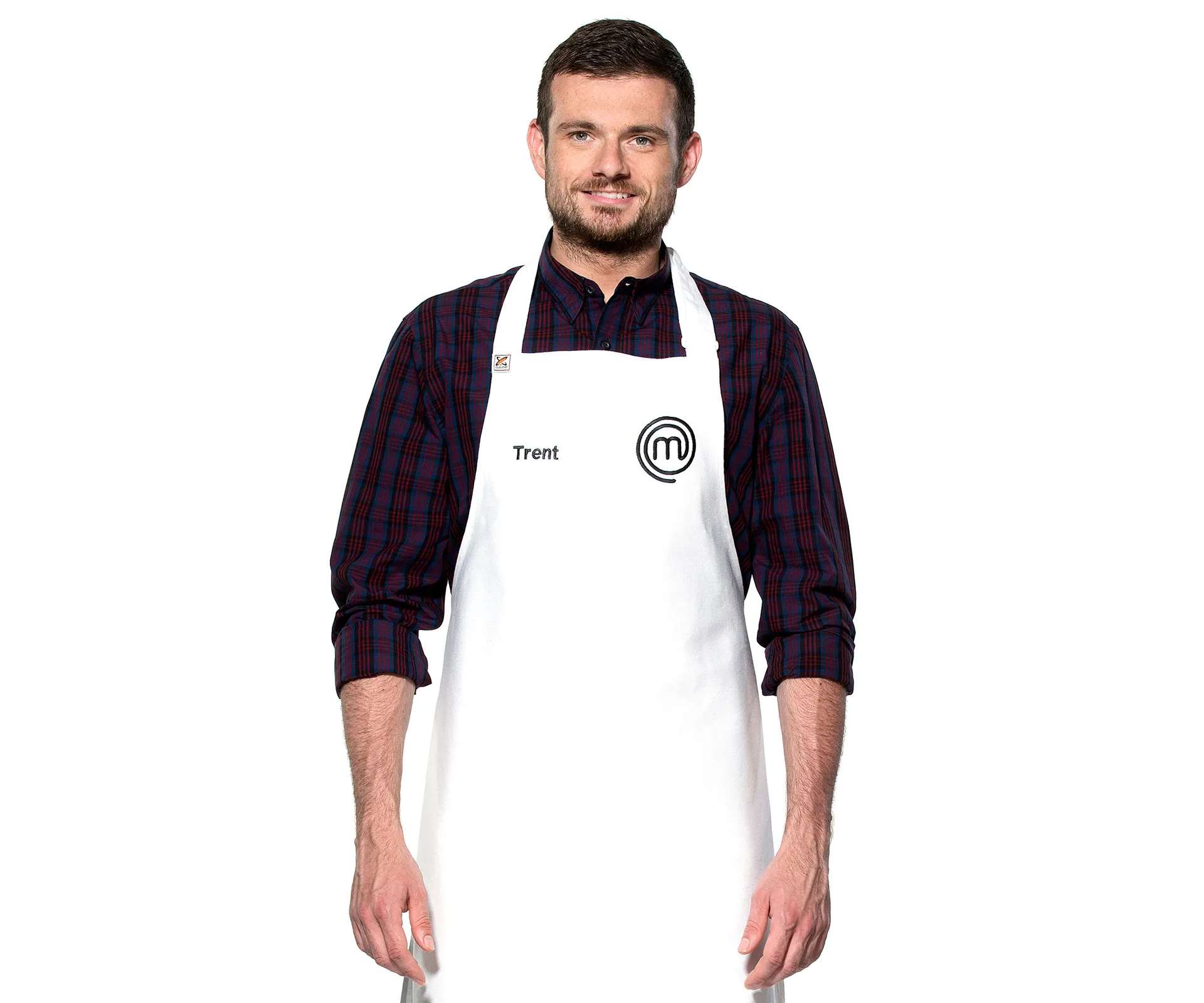 Trent from MasterChef Australia wearing a white apron with a MasterChef logo and a plaid shirt, smiling.