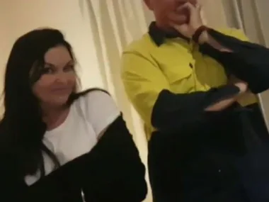 Two people indoors; one woman in a white top smiles at the camera, and a man in a yellow shirt has a hand on his chin.