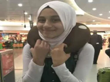 Young girl in a hijab at an airport, smiling with a neck pillow.